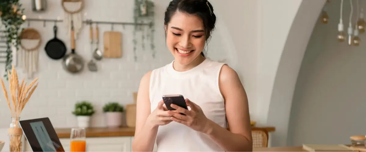 Lifestyle Moment - Woman Smiling at Phone: A happy resident in her Hillhaven kitchen, smiling as she reads on her phone, showcasing the warm and community-oriented lifestyle at Hillhaven.