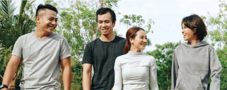 Group of People Walking Outdoors - A group of four people, casually dressed, walking and smiling outdoors in a lush green setting, showing a sense of community that's possible at Hillhaven.